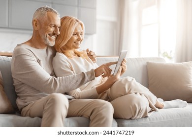 Elderly couple sharing a joyful moment while looking at a tablet together, seated in a comfortable living room - Powered by Shutterstock