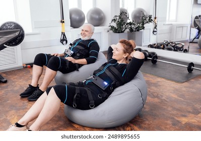 Elderly couple sits on massage chairs while waiting for ems workout in gym or fitness club. Senior greyhaired man and woman in ems suits getting ready for electrical muscle stimulation training. - Powered by Shutterstock