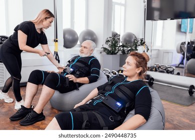 Elderly couple sits on massage chairs while waiting for ems workout in gym. Young female trainer using remote control sets necessary parameters on electrical muscle stimulation suit of senior man. - Powered by Shutterstock