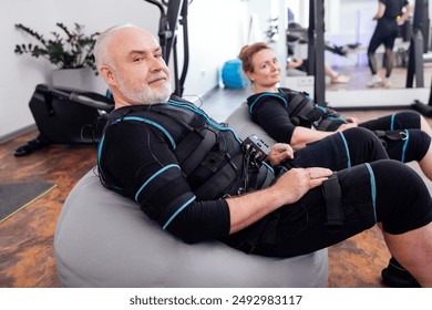 Elderly couple sits on massage chairs while waiting for ems workout in gym or fitness club. Senior greyhaired man and woman in ems suits getting ready for electrical muscle stimulation training. - Powered by Shutterstock