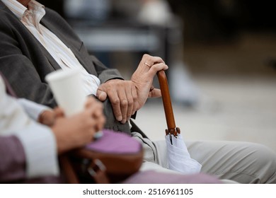 An elderly couple sits together, holding hands. One holds a wooden cane and a folded white umbrella. The focus is on their hands, symbolizing closeness, aging, and a peaceful moment of togetherness. - Powered by Shutterstock