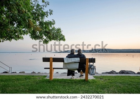 An elderly couple sitting on a bench, looking at Lake Balaton in the summer twilight. The man is embracing the woman's shoulder. They have a drying towel and a bag with them - front horizontal view