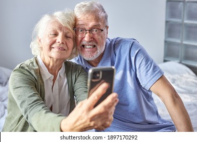 elderly couple sitting on a couch taking photo on smartphone, posing at phone's camera, enjoying time at weekends. family, technology, age and people concept