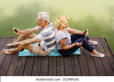 Elderly Couple Sitting At Pier. Old Man Holding Hands Mobile Phone While Sitting Back To Back Senior Woman At The Shore.
Active Retired Woman Holding Hands Digital Tablet.