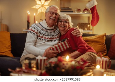 Elderly couple smiling and hugging on the sofa while grandpa is holding a christmas present his wife got for him - Powered by Shutterstock