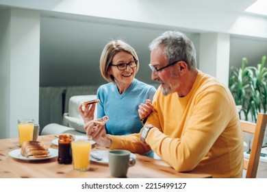 Elderly couple spending weekend day at home while talking in a bright room with modern interior, enjoying moment and eating breakfast together. Eating french toast. Focus on a smiling mature woman. - Powered by Shutterstock