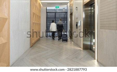 An elderly couple stepped out of the elevator in the hotel lobby. They were casually dressed and carrying suitcase, having checked out after traveling.