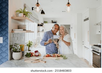 Elderly couple taking a selfie while cooking in a kitchen. Wine and alcohol drink concept. Retired couple standing in kitchen drinking wine. Senior couple in love in their home. - Powered by Shutterstock