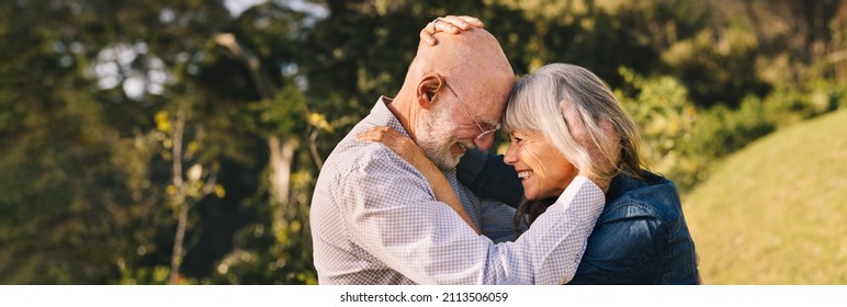 Elderly Couple Touching Their Heads Together While Standing Outdoors. Happy Senior Couple Sharing A Romantic Moment In A Park. Cheerful Mature Couple Expressing Their Love And Affection.