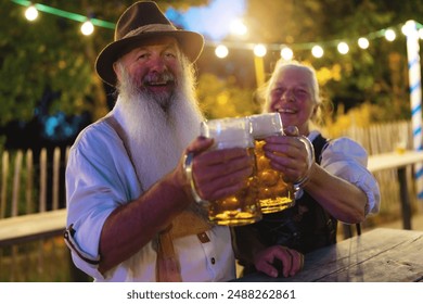 An elderly couple in traditional Bavarian tracht raising beer mugs and smiling at Oktoberfest or dult in beer garden with festive lights in the background - Powered by Shutterstock