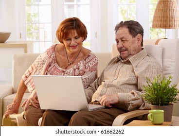 Elderly Couple Using Laptop Computer At Home, Looking At Screen, Smiling.?