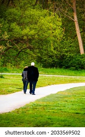 An elderly couple walk a long a path in a park, holding each other. Couple is walking away from view with arm around her.