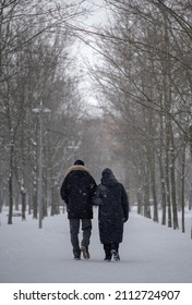 An Elderly Couple Walking Away Down The Road. Pensioners Go Into The Depths Of The Park. Elderly People Walk By The Hand In The Winter Park.