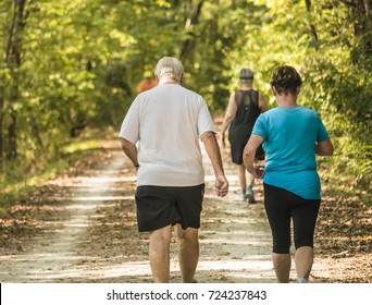 Elderly Couple Walking On A Trail For Exercise; Woods On The Sides Of The Trail; Man Is Overweight