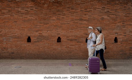 Elderly couple walking with suitcase along ancient brick wall in Chiang Mai, Thailand. Woman in floral shirt, man in hat. Scene of senior tourists traveling and exploring local heritage. - Powered by Shutterstock