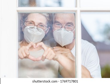 Elderly couple wearing protective face masks watch through their home window and show heart sign during the coronavirus epidemic - Powered by Shutterstock