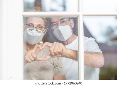 Elderly couple wearing protective face masks watch through their home window and show heart sign during the coronavirus epidemic - Powered by Shutterstock