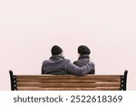 An elderly couple in winter coats sit on a bench isolated on pastel pink background. The elderly couple enjoys a winter moment. The elderly couple shares warmth on a cold winter day. Winter love.