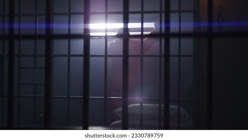 Elderly criminal in orange uniform sits in jail cell, stands up and looks on window with bars. Guilty inmate in detention center or correctional facility. Prisoner serves imprisonment term.