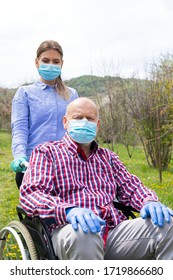 Elderly Disabled Man With Mask Sitting In Wheelchair, Assisted By Young Female Caregiver Outdoors