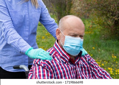 Elderly Disabled Man With Mask Sitting In Wheelchair, Assisted By Young Female Caregiver Outdoors