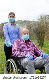 Ältere behinderte Menschen mit Maske, die im Rollstuhl sitzen, unterstützt von einer jungen weiblichen Betreuerin im Freien