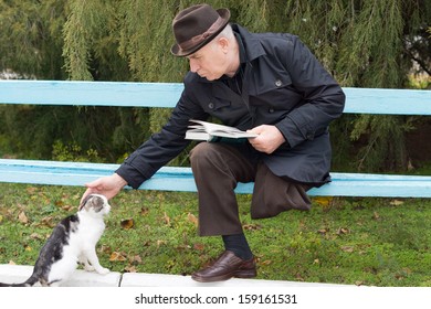 Elderly Disabled Man With One Leg Amputated Sitting On A Park Bench With His Book Reaching Down And Stroking A Cat