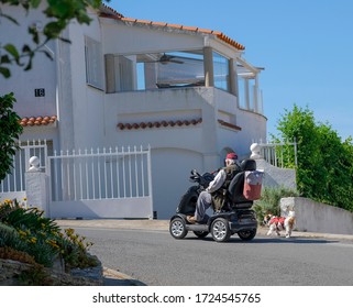 Elderly Disabled Person Or Senior With A Dog On Electric Wheelchair Or A Mobility Scooter Driving On Road. An Elderly Person Is Always Together With His Pet. Modern Life In Europe For Disabilities.