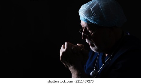 An Elderly Doctor With A Stethoscope And A Cap On A Black Background In Profile, Holds His Hands Near His Face, Prays, Close-up, Space For Text, Banner