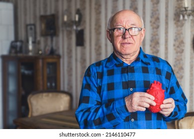 Elderly European Bald Senior Man In Blue Flannel Shirt Holding Artificial Fake Heart Prop In Front Of His Chest. Old-fashioned Interior In The Background. Healthcare Concept. High Quality Photo