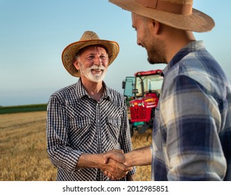 Elderly farmer shaking hands with young male farmer standing in harvested field with tractor in background.
