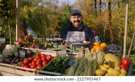 Elderly farmer stands at the stall with fresh colorful fruits and vegetables, looks at camera. Weekend shopping at local farmers market outside. Vegetarian, organic and healthy food. Agriculture.