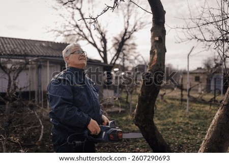 Elderly farmer wearing protective glasses and holding chainsaw while pruning fruit trees in his orchard during winter