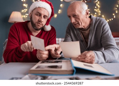 An elderly father looks at old photos with his adult son and shows pictures of him when he was young. The concept of family traditions. Copy space - Powered by Shutterstock