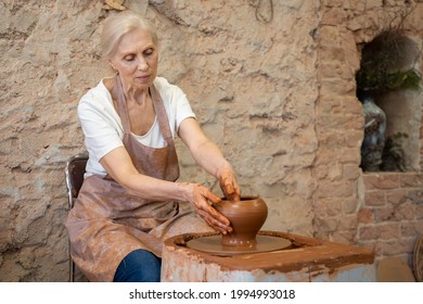 Elderly female potter working on pottery wheel while sitting in her workshop - Powered by Shutterstock