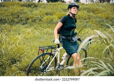 Elderly Female Relaxing After Riding Bicycle In Green Nature. Senior Woman Cyclist In Black Sportswear And Helmet In Green Meadow.  Copy Space