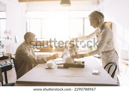 elderly female standing and pouring coffee from kettle into teapot at table with senior male blogger sitting and looking at screen of laptop while collecting blog information from internet at home
