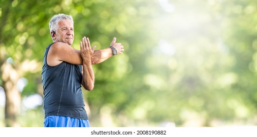 Elderly fit man stretches his arm on a summer day outdoors, wearing watches and earphones.