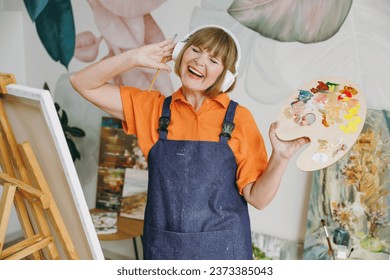 Elderly fun artist woman 50 years old wear casual clothes stand near easel with painting artwork paint listen to music in headphones spend free spare time in living room indoor. Leisure hobby concept - Powered by Shutterstock