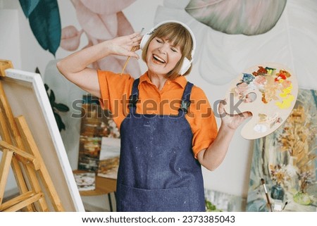Elderly fun artist woman 50 years old wear casual clothes stand near easel with painting artwork paint listen to music in headphones spend free spare time in living room indoor. Leisure hobby concept