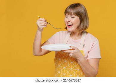 Elderly Fun Happy Cooker Housekeeper Housewife Woman 50s In Orange Apron Hold Empty Plate Spoon Try Meal Eat Isolated Plain On Yellow Background Studio Portrait. People Household Lifestyle Concept.