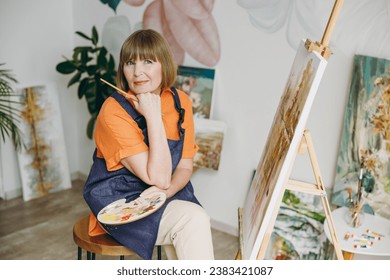 Elderly fun minded artist woman 50 years old wears casual clothes sit near easel with painting artwork sit paint hold palette brush spend free spare time in living room indoor. Leisure hobby concept - Powered by Shutterstock