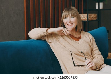 Elderly fun minded happy woman 50s years old wears casual clothes sits on blue sofa look aside hold in hand glasses stay at home flat rest relax spend free spare time in living room indoor grey wall - Powered by Shutterstock