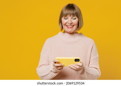 Elderly Gambling Woman 50s Wears Pink Casual Knitted Sweater Using Play Racing App On Mobile Cell Phone Hold Gadget Smartphone For Pc Video Games Isolated On Plain Yellow Background Studio Portrait.