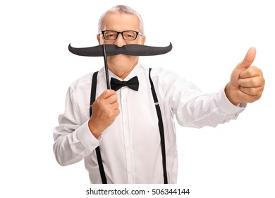 Elderly Gentleman In A Bow Tie And Suspenders Posing With A Big Fake Moustache And Giving A Thumb Up Isolated On White Background