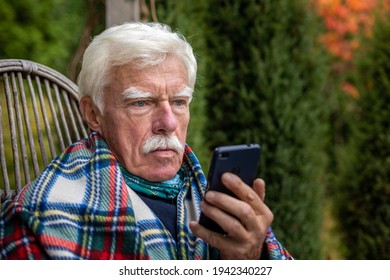 An Elderly Gentleman Is Having A Phone Call. Senior Man Sitting In The Rocking Chair During His Smartphone Conversation. Mature Man Using His Cellular On The Veranda.