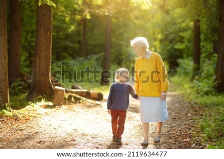 Elderly grandmother and her little grandchild walking together in sunny summer park. Friendship of grandma and grandson. Two generations of family.
