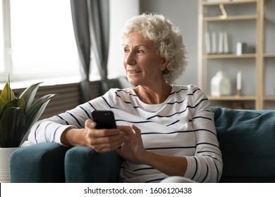 Elderly grandmother resting on couch holding in hands smart phone distracted from chatting with grown up children looking out the window enjoy day feels healthy. Older generation using gadgets concept