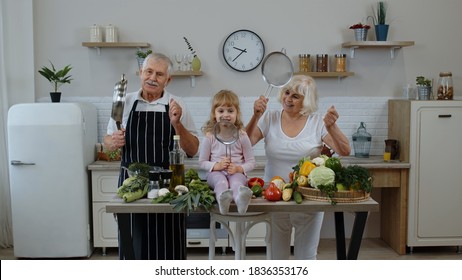 Elderly Grandparents Dancing With Grandchild Girl While Cooking Together In Kitchen At Home. Senior Woman And Man Making A Funny Dance With Strainer And Vegetables. Healthy Mature Family