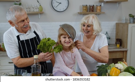 Elderly Grandparents Dancing With Grandchild Girl While Cooking Together In Kitchen At Home. Senior Woman And Man Making A Funny Dance With Strainer And Vegetables. Healthy Mature Family